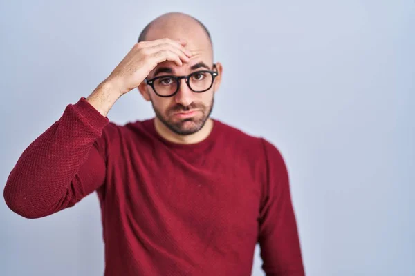 Young bald man with beard standing over white background wearing glasses worried and stressed about a problem with hand on forehead, nervous and anxious for crisis