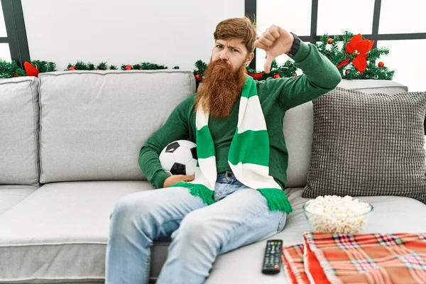 Redhead man with long beard football hooligan holding ball sitting on the sofa with angry face, negative sign showing dislike with thumbs down, rejection concept