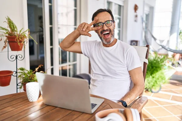 Middle age man using computer laptop at home doing peace symbol with fingers over face, smiling cheerful showing victory