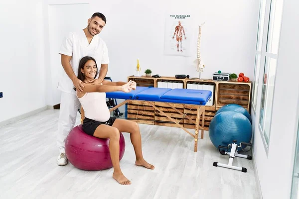 Latin man and woman wearing physiotherapist uniform having pregnancy rehab session using fit ball at physiotherapy clinic