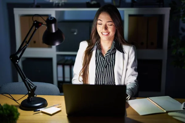 Young brunette woman working at the office at night with laptop winking looking at the camera with sexy expression, cheerful and happy face.