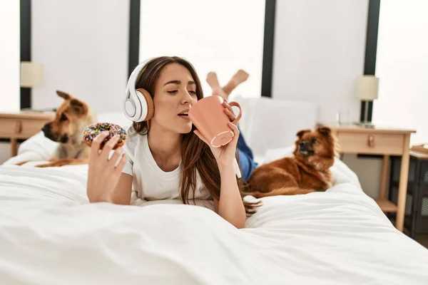 Young hispanic woman having breakfast and listening to music lying on bed with dogs at bedroom