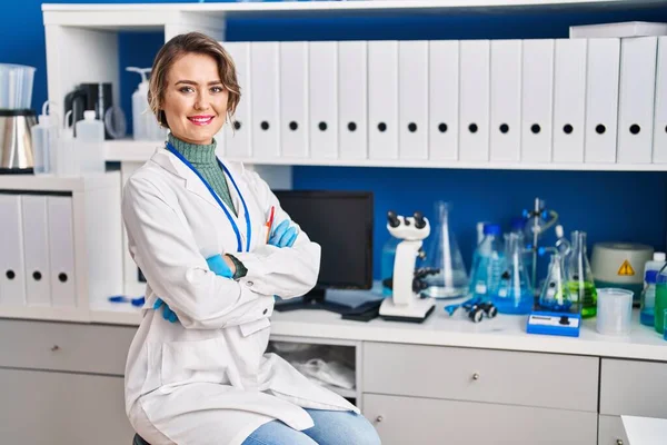 Young woman scientist smiling confident sitting with arms crossed gesture at laboratory