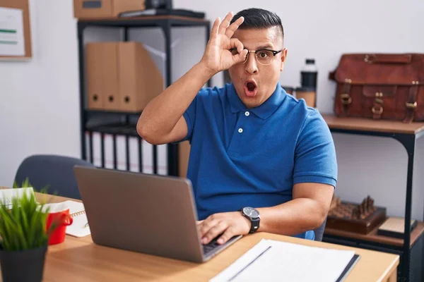 Young hispanic man working at the office with laptop doing ok gesture shocked with surprised face, eye looking through fingers. unbelieving expression.