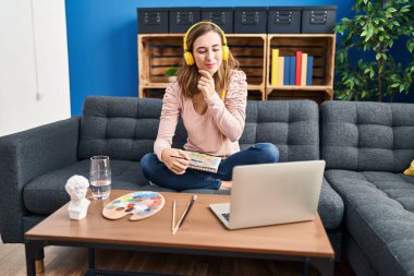Young beautiful woman practicing painting looking at online tutorial serious face thinking about question with hand on chin, thoughtful about confusing idea 