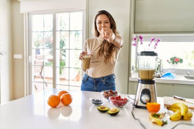 Güzel, genç, esmer bir kadın mutfakta bir bardak smoothie içerek kamerayı işaret ediyor ve sen kendine güvenen ciddi bir jest yapıyorsun. 
