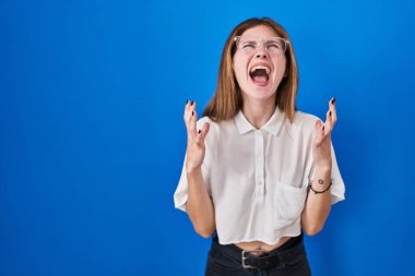 Güzel bir kadın mavi arka planda duruyor deli gibi bağırıyor ve bağırıyor agresif bir ifade ve kollar havada. hüsran kavramı. 