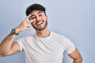 Hispanic man with beard standing over white background doing peace symbol with fingers over face, smiling cheerful showing victory 