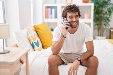 Genç İspanyol adam akıllı telefonda konuşuyor. Yatak odasında oturuyor.
