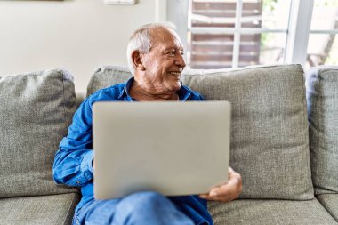Evinin oturma odasındaki kanepede oturan gri saçlı yaşlı adam bilgisayar laptopu kullanıyor.