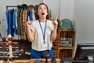 Beautiful middle age hispanic woman working as manager at retail boutique amazed and surprised looking up and pointing with fingers and raised arms. 