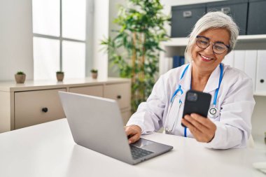 Klinikte doktor üniforması giyen orta yaşlı bir kadın akıllı telefon kullanıyor.