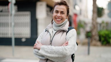 Orta yaşlı İspanyol kadın gülümsüyor. Kollarını kavuşturup caddede duruyor.