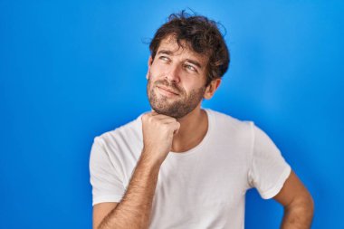 Mavi arka planda dikilen İspanyol genç adam elleri çenesinde soru sorarken, düşünceli bir ifade takınıyor. Düşünceli bir yüzle gülümsüyor. şüphe kavramı. 