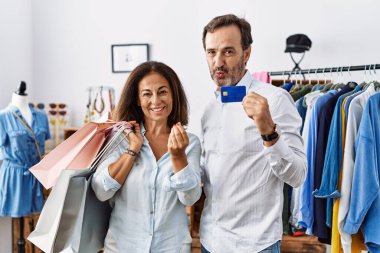 Hispanic middle age couple holding shopping bags and credit card doing money gesture with hands, asking for salary payment, millionaire business 