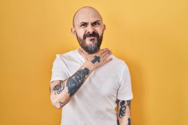 Young hispanic man with tattoos standing over yellow background cutting throat with hand as knife, threaten aggression with furious violence 