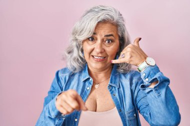 Middle age woman with grey hair standing over pink background smiling doing talking on the telephone gesture and pointing to you. call me. 