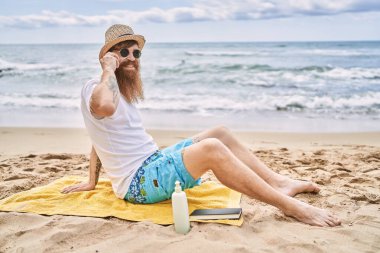 Genç kızıl saçlı adam kumsalda havlunun üzerinde oturmuş akıllı telefondan konuşuyor..