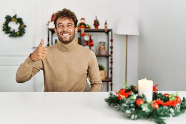 Noel süslemesi masasında oturan sakallı yakışıklı genç adam elleriyle mutlu baş parmaklar hareketi yapıyor. Kameraya bakıp başarıyı gösteren ifade. 
