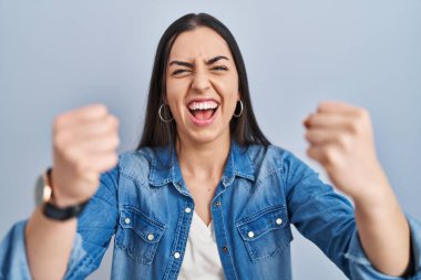 Mavi arka planda öfkeli ve kızgın duran İspanyol kadın öfkeyle bağırırken yumruklarını kaldırıyor ve sinirleniyor. öfke ve agresif konsept. 