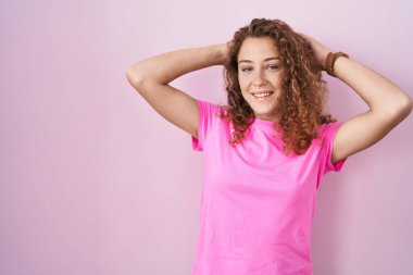 Genç beyaz kadın pembe arka planda duruyor rahatlıyor ve esniyor, kolları ve elleri başının ve boynunun arkasında mutlu bir şekilde gülümsüyor. 