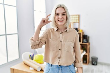 Young beautiful caucasian woman at construction office smiling and confident gesturing with hand doing small size sign with fingers looking and the camera. measure concept. 