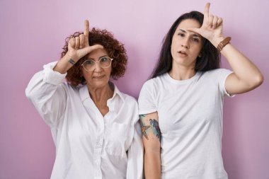 İspanyol anne ve kızı pembe arka plan üzerine gündelik beyaz t-shirt giyiyorlar. Alnında parmakları olan insanlarla dalga geçiyorlar. Ezik jestler yapıp, alay ediyorlar.. 