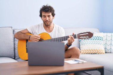 Latin kökenli genç bir adam evdeki kanepede oturup online gitar dersi alıyor.