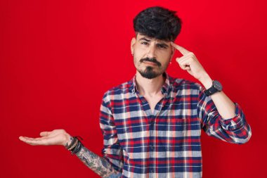 Kırmızı arka planda duran sakallı genç İspanyol adam. Avuç içi açık fotokopi alanı ve alnını göstermekten rahatsız ve kafası karışık. Bunu bir düşün.. 