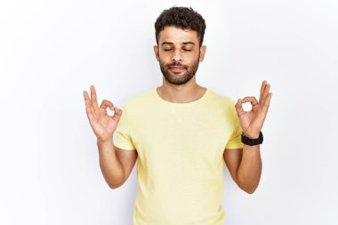 Arap genç adam izole edilmiş arka planda duruyor rahatla ve gözleri kapalı gülümse parmaklarıyla meditasyon hareketi yapıyor. yoga konsepti. 