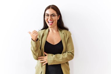 Genç esmer kadın izole edilmiş arka planda gülümsüyor, gülümsüyor ve başparmağıyla yan tarafı işaret ediyor.. 