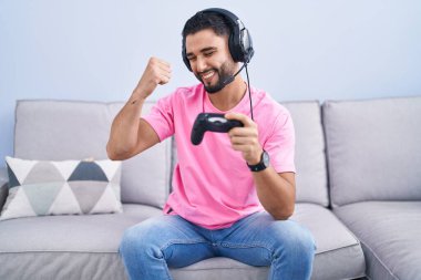 Kanepede oturmuş video oyunu oynayan İspanyol genç adam çok mutlu ve heyecanlı bir şekilde kollarını kaldırarak, gülümseyerek ve başarı çığlıkları atarak kazanan hareketi yapıyor. kutlama konsepti. 