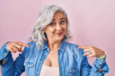Middle age woman with grey hair standing over pink background looking confident with smile on face, pointing oneself with fingers proud and happy. 