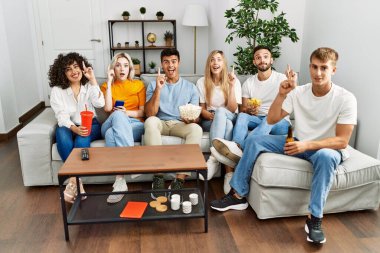 Group of people eating snack sitting on the sofa at home surprised with an idea or question pointing finger with happy face, number one 