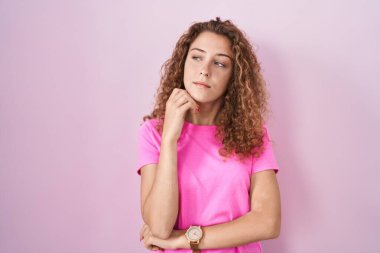 Pembe arka planda, elleri çenesinde soru sormayı düşünen beyaz bir kadın. Düşünceli bir ifade. Düşünceli bir yüzle gülümsüyor. şüphe kavramı. 