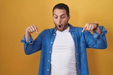 Hispanic man standing over yellow background pointing down with fingers showing advertisement, surprised face and open mouth 