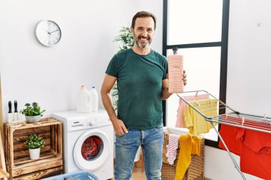 Middle age man with beard doing laundry holding detergent bottle looking positive and happy standing and smiling with a confident smile showing teeth 
