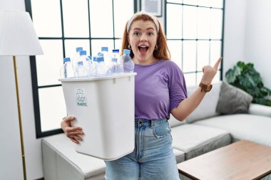 Geri dönüşüm sepetini elinde tutan genç kızıl saçlı kadın yüzünde kocaman bir gülümseme olan plastik şişelerle kameraya el ile işaret ediyor.. 