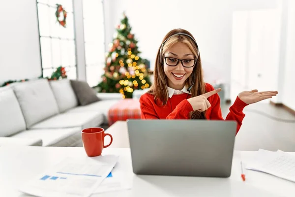 年轻的高加索女孩坐在桌子上 用笔记本电脑在圣诞树旁工作 一边手牵着手 一边指指点点着相机 一边惊讶地微笑着 — 图库照片