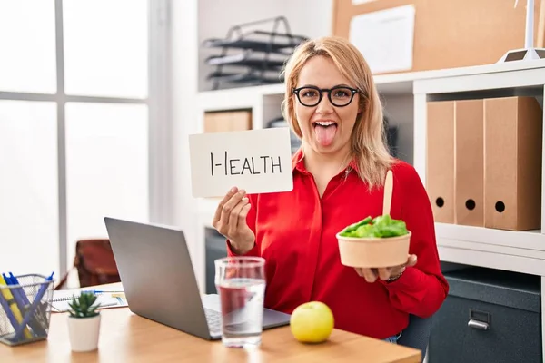 在办公室工作的金发女人吃着健康的食物 伸出舌头 带着滑稽的表情高兴极了 — 图库照片