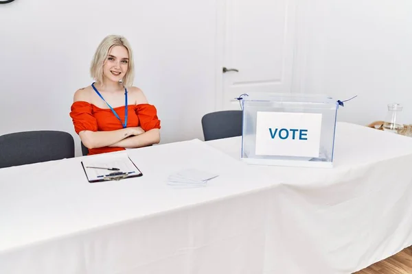 Young caucasian woman at political election sitting by ballot happy face smiling with crossed arms looking at the camera. positive person.