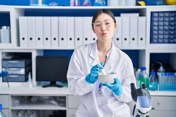 Chinese young woman working at scientist laboratory mixing looking at the camera blowing a kiss being lovely and sexy. love expression.