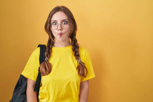 Young caucasian woman wearing student backpack over yellow background making fish face with lips, crazy and comical gesture. funny expression.