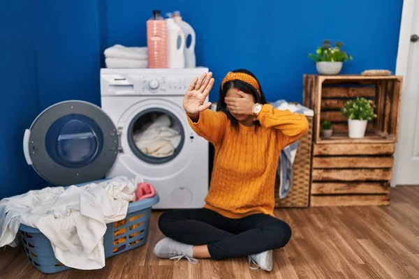 Young hispanic woman doing laundry covering eyes with hands and doing stop gesture with sad and fear expression. embarrassed and negative concept.