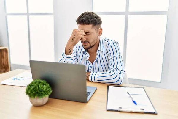 年轻英俊的男人留着胡子 在办公室里用电脑笔记本电脑工作 疲惫的揉揉鼻子和眼睛 感觉疲劳和头痛 压力和挫败感概念 — 图库照片