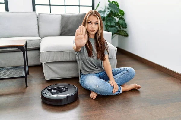 Young caucasian woman sitting at home by vacuum robot with open hand doing stop sign with serious and confident expression, defense gesture