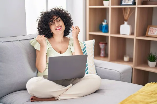 Young brunette woman with curly hair using laptop sitting on the sofa at home very happy and excited doing winner gesture with arms raised, smiling and screaming for success. celebration concept.