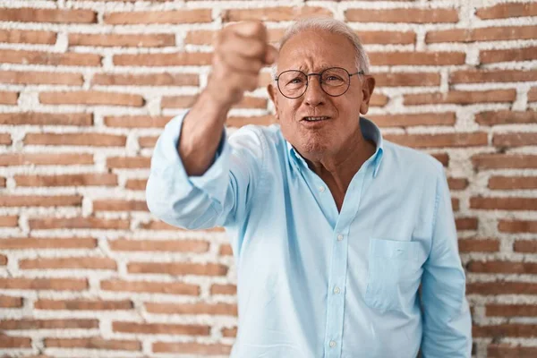 Senior man with grey hair standing over bricks wall angry and mad raising fist frustrated and furious while shouting with anger. rage and aggressive concept.