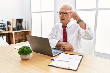 Senior man working at the office using computer laptop shooting and killing oneself pointing hand and fingers to head like gun, suicide gesture. 