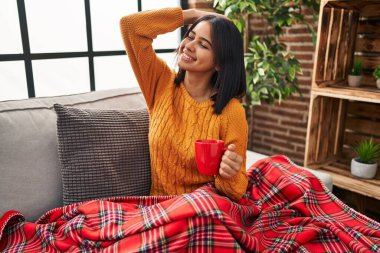 Genç İspanyol kadın kanepede oturup kahvesini yudumlarken gülümseyerek saçlarına dokunup el hareketleriyle çekici ve şık bir poz veriyor. 